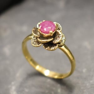 Könnte beinhalten: Ein goldfarbener Ring mit floralem Design. Der Ring hat einen zentralen, runden, rosa Edelstein, umgeben von detaillierten Blütenblättern. Das Band ist glatt und schlank, wodurch ein elegantes und feminines Schmuckstück entsteht.