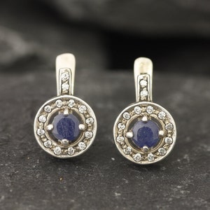 May include: A pair of silver earrings with a blue gemstone surrounded by a halo of smaller white gemstones.