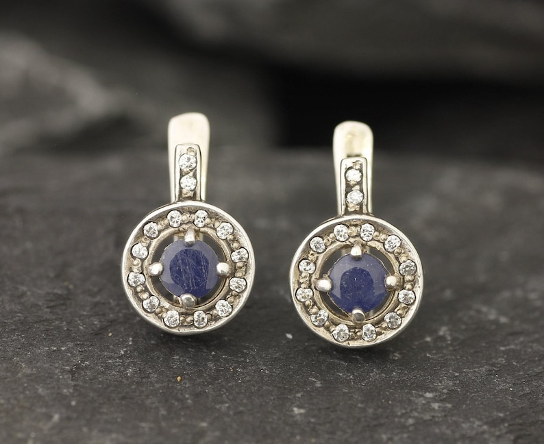 May include: A pair of silver earrings with a blue gemstone surrounded by a halo of smaller white gemstones.
