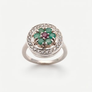 Puede incluir: Un anillo de plata con un diseño floral. El centro del anillo tiene una piedra preciosa de color rojo oscuro rodeada de piedras preciosas verdes. El anillo exterior está incrustado con pequeñas piedras preciosas claras.