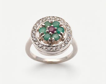 Puede incluir: Un anillo de plata con un diseño floral. El centro del anillo tiene una piedra preciosa de color rojo oscuro rodeada de piedras preciosas verdes. El anillo exterior está incrustado con pequeñas piedras preciosas claras.