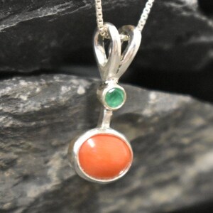 Puede incluir: Un colgante de plata con una piedra redonda de coral naranja y una peque&ntilde;a piedra esmeralda verde.