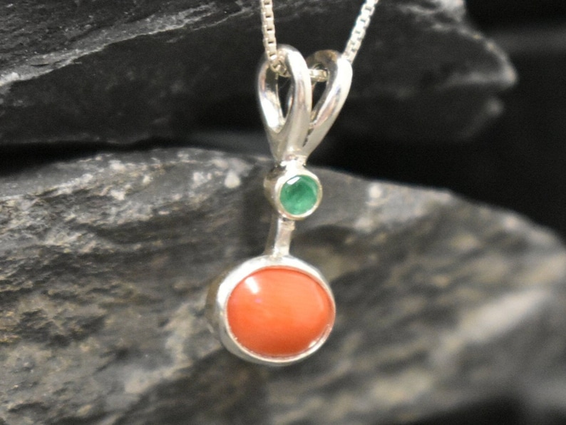 Puede incluir: Un colgante de plata con una piedra redonda de coral naranja y una peque&ntilde;a piedra esmeralda verde.