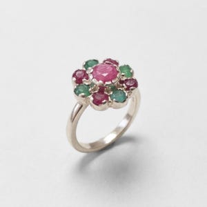May include: A silver ring featuring a central round faceted pink ruby surrounded by a cluster of alternating smaller round faceted pink rubies and green emeralds in a floral arrangement, set on a smooth, polished silver band.