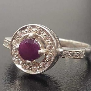 May include: A silver ring featuring a round, purple gemstone encircled by smaller white gemstones. The ring has a plain band.