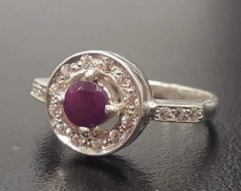 May include: A silver ring featuring a round, purple gemstone encircled by smaller white gemstones. The ring has a plain band.
