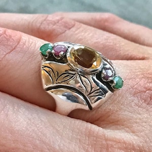 Könnte beinhalten: Ein silberner Ring mit einem großen, facettierten, gelben Edelstein, der von kleineren grünen und rosa Edelsteinen umgeben ist. Der Ring hat ein detailliertes Blumenmuster.