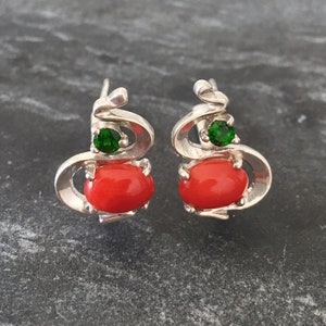 Pu&ograve; includere: Un paio di orecchini d'argento con un design in corallo rosso e una pietra preziosa verde. Gli orecchini hanno la forma di una spirale.