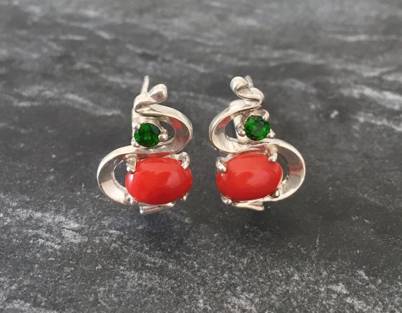 Pu&ograve; includere: Un paio di orecchini d'argento con un design in corallo rosso e una pietra preziosa verde. Gli orecchini hanno la forma di una spirale.