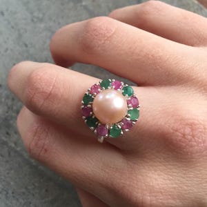 May include: A silver ring featuring a large, light pink pearl center, surrounded by a halo of alternating green and red gemstones. The ring is set against a neutral background, showcasing the jewelry's design.