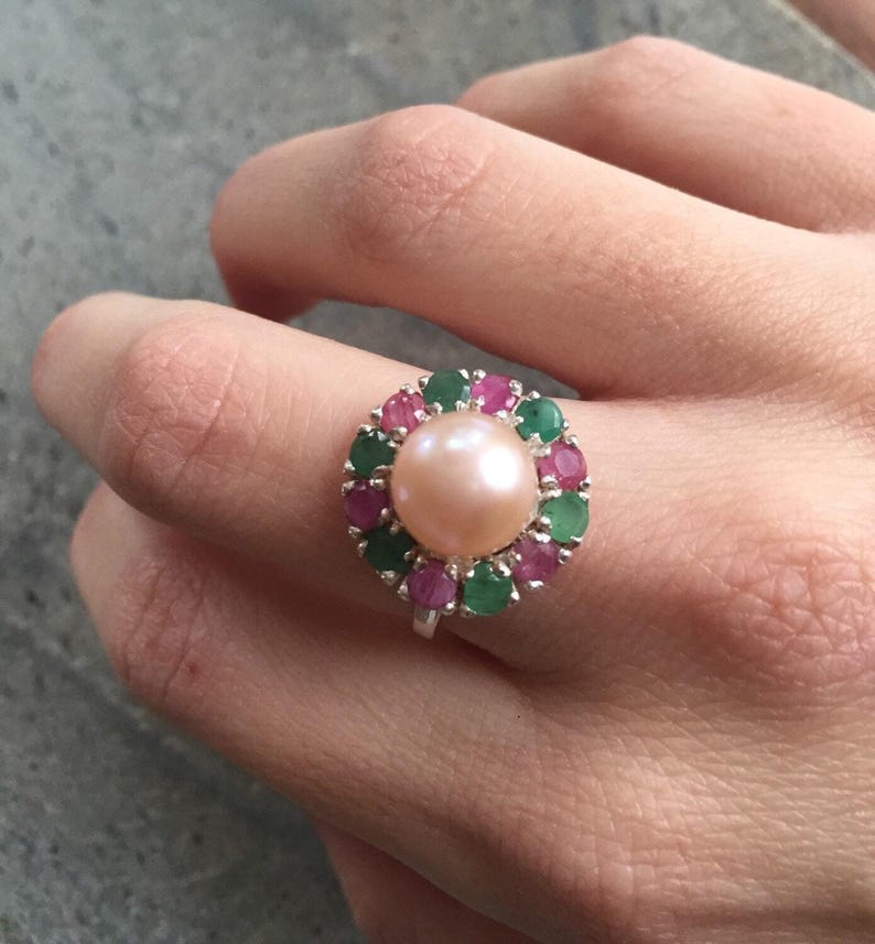 May include: A silver ring featuring a large, light pink pearl center, surrounded by a halo of alternating green and red gemstones. The ring is set against a neutral background, showcasing the jewelry's design.