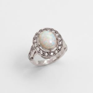 May include: A silver-toned ring featuring a central oval-shaped iridescent opal cabochon. The opal is surrounded by a halo of small, clear, round-cut stones set into the metal frame, with additional small stones embedded along the ring band.