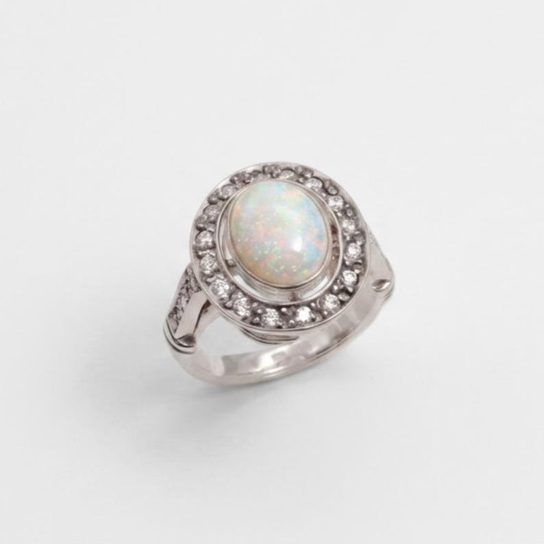 May include: A silver-toned ring featuring a central oval-shaped iridescent opal cabochon. The opal is surrounded by a halo of small, clear, round-cut stones set into the metal frame, with additional small stones embedded along the ring band.