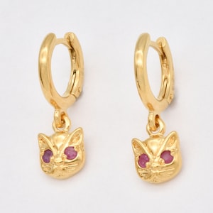 Puede incluir: Pendientes de aro de oro con un dije de gato colgante. El dije de gato tiene ojos de piedras preciosas rosas.