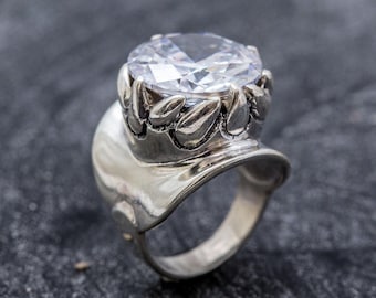 May include: A silver ring with a large, clear, round gemstone set in a crown-like design. The ring band is wide and has a textured, organic shape.