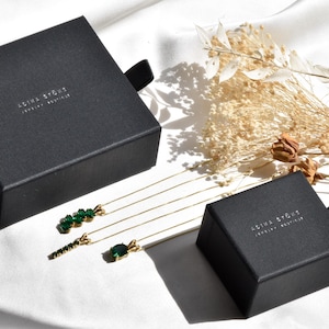 Puede incluir: Tres collares dorados con colgantes de piedras preciosas verdes se muestran junto a dos cajas de joyer&iacute;a negras con la etiqueta ADINA STONE JEWELRY BOUTIQUE. Los collares presentan diferentes estilos de colgantes, incluyendo un grupo, una barra y una piedra ovalada.