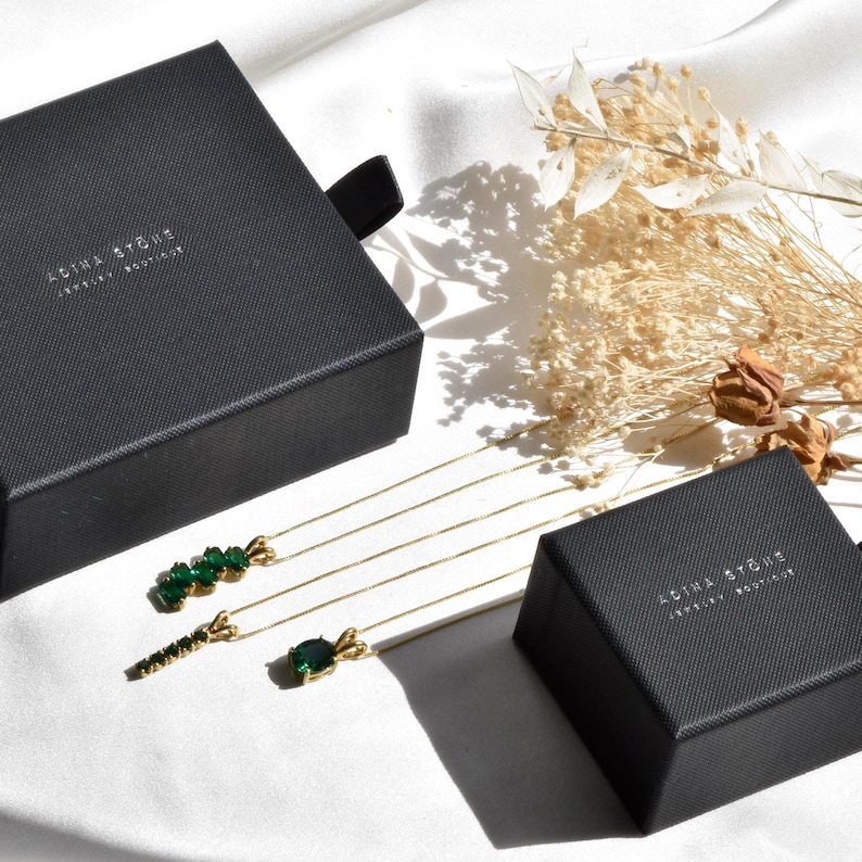 Puede incluir: Tres collares dorados con colgantes de piedras preciosas verdes se muestran junto a dos cajas de joyer&iacute;a negras con la etiqueta ADINA STONE JEWELRY BOUTIQUE. Los collares presentan diferentes estilos de colgantes, incluyendo un grupo, una barra y una piedra ovalada.