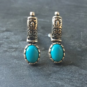 May include: A pair of silver earrings with turquoise stones. The earrings have a decorative design on the top part and a round turquoise stone at the bottom.