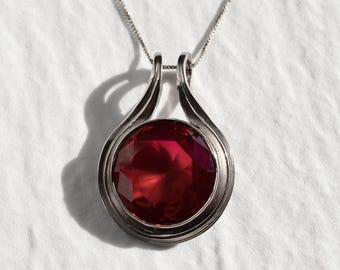 May include: A silver necklace with a red gemstone pendant. The pendant is round and has a faceted design. The necklace is on a white background.