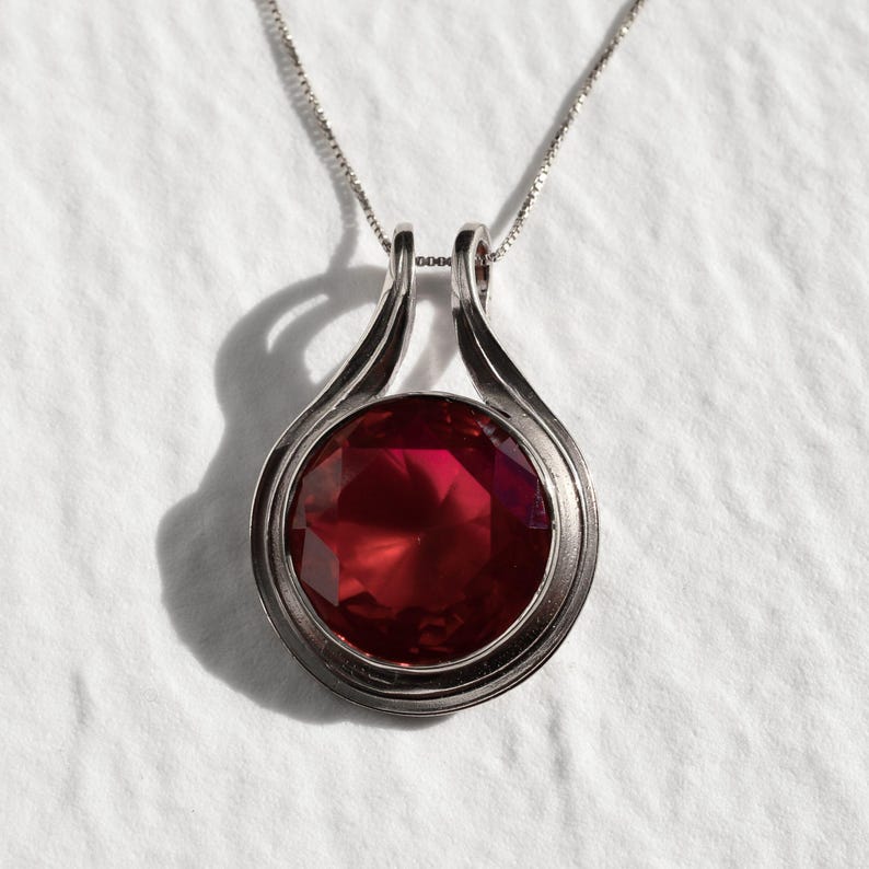 May include: A silver necklace with a red gemstone pendant. The pendant is round and has a faceted design. The necklace is on a white background.