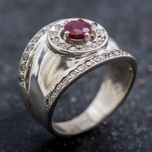 May include: A silver ring featuring a central, oval, deep red gemstone encircled by small, clear stones. The ring's band is also embellished with small, clear stones, and has a distinctive, swirling design.