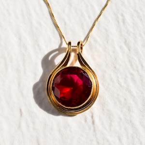 Pode incluir: Um colar de ouro com um pingente que apresenta uma grande gema vermelha redonda engastada em uma moldura de ouro.