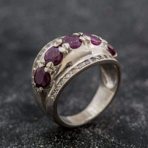 May include: A silver-toned ring featuring a row of five deep red oval-cut rubies set horizontally across the centre. The band is accented with a line of small, clear pavé-set stones along the upper and lower edges of the central ruby setting.