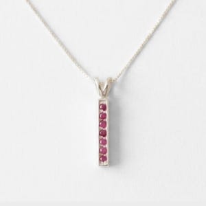 Può includere: Una collana color argento con un pendente a barra verticale incastonato con sei piccole pietre preziose rotonde di un rosso rosato intenso disposte in colonna. Il pendente è appeso a una sottile e delicata catenina color argento.