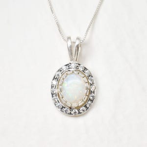 May include: A silver pendant necklace featuring a central oval-shaped iridescent opal stone surrounded by a halo of small, clear, round-cut stones. The pendant is suspended from a delicate silver box chain, creating a classic and elegant jewelry piece.