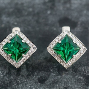 May include: A pair of silver earrings featuring a square emerald green gemstone set within a halo of smaller clear gemstones.