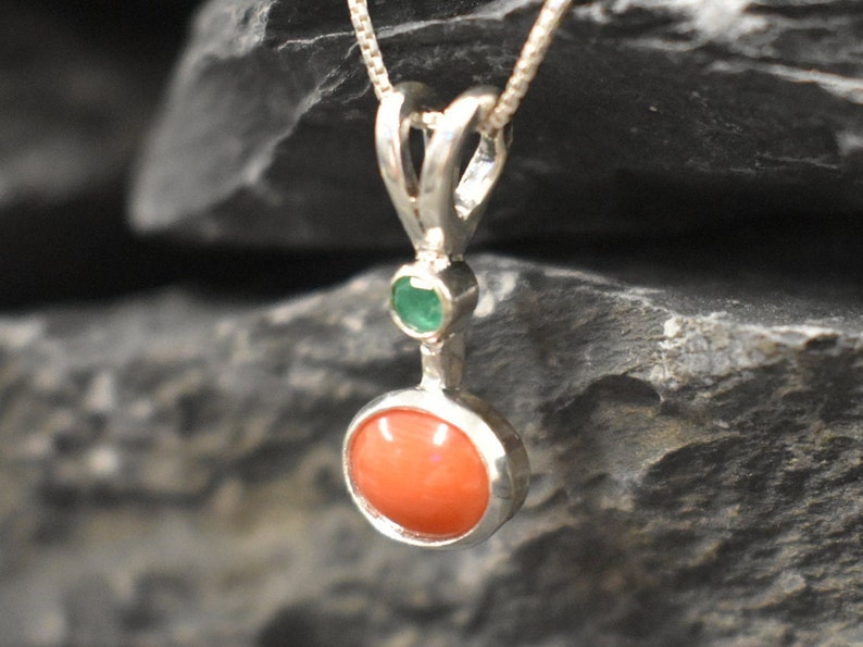 Puede incluir: Un collar con un colgante de plata, adornado con una piedra redonda de coral naranja y una peque&ntilde;a piedra redonda de esmeralda verde.