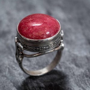 Puede incluir: Un anillo de plata con una gran piedra preciosa redonda de color rojo engastada en un engaste de bisel. El anillo tiene detalles intrincados alrededor de la piedra preciosa.