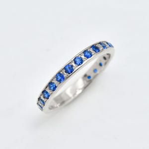 Op de afbeelding: Een zilverkleurige metalen ring met een rij kleine, ronde, levendig blauwe edelstenen die langs de bovenste helft van de band zijn gezet. De ring heeft een gepolijste afwerking en een slank profiel met een consistente rij blauwe stenen.