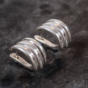May include: A pair of silver-colored, ribbed hoop earrings. Each earring has a curved, semi-circular design with multiple parallel ridges. The earrings are set against a dark, textured background, highlighting their metallic sheen.