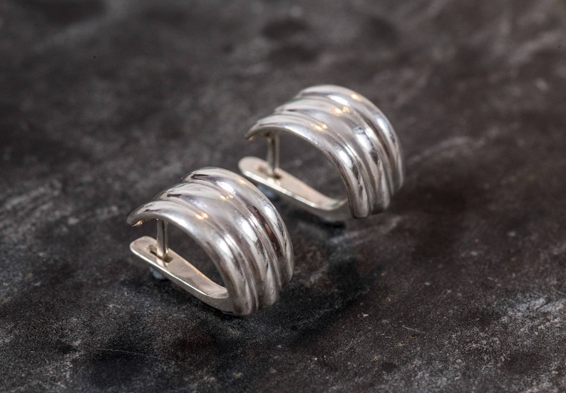 May include: A pair of silver-colored, ribbed hoop earrings. Each earring has a curved, semi-circular design with multiple parallel ridges. The earrings are set against a dark, textured background, highlighting their metallic sheen.