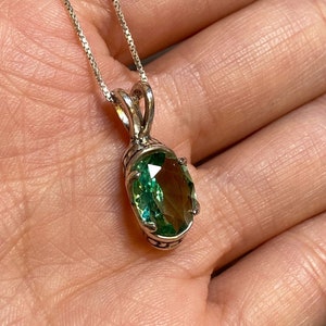 Puede incluir: Un collar de cadena de plata con un colgante de piedra preciosa ovalada verde. El colgante tiene un engaste de plata detallado.
