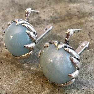 Op de afbeelding: Een paar zilveren oorbellen met lichtblauwe edelsteen cabochons, gezet in een zilveren klauw setting.