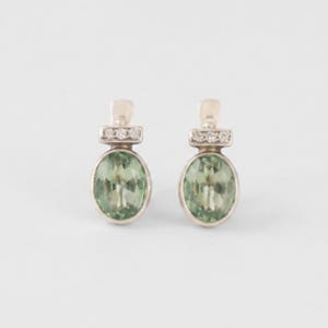 Puede incluir: Un par de pendientes con piedras preciosas de color verde claro en corte ovalado engastadas en metal plateado. Cada pendiente tiene una barra horizontal sobre la piedra ovalada, decorada con tres pequeñas piedras claras de corte redondo.