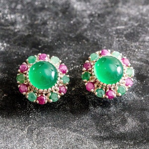 Op de afbeelding: Een paar oorstekers met een groot, rond, heldergroen edelsteen centrum, omringd door kleinere, afwisselend groene en roze edelstenen. De oorbellen hebben een zilverkleurige metalen zetting.