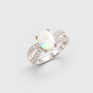 May include: A silver-toned ring featuring a central oval-shaped opal gemstone with iridescent flashes of color. The band has a split-shank design, with each side encrusted with small, clear, round-cut stones set in a polished metal finish.