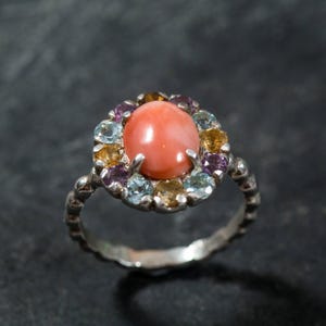 Puede incluir: Un anillo de plata con una piedra preciosa de coral rosa rodeada por un halo de piedras preciosas m&aacute;s peque&ntilde;as en tonos azules, amarillos y morados.
