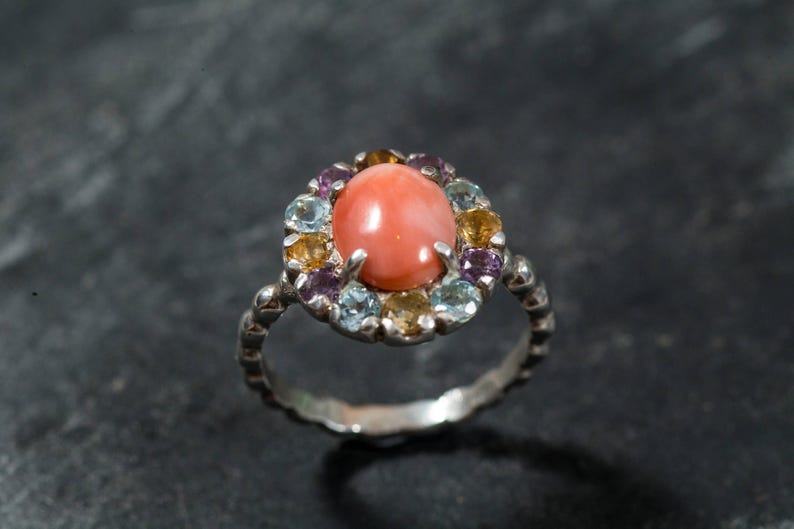 Puede incluir: Un anillo de plata con una piedra preciosa de coral rosa rodeada por un halo de piedras preciosas m&aacute;s peque&ntilde;as en tonos azules, amarillos y morados.