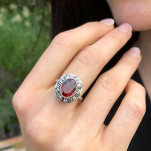 Puede incluir: Un anillo de plata con una gran piedra roja ovalada rodeada de piedras blancas más pequeñas.