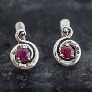 May include: A pair of silver earrings with a swirling design and a small, round, red gemstone in the center of each earring.