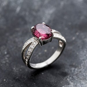 Pu&ograve; includere: Un anello d'argento con una grande gemma ovale rosa intenso. La fascia si divide in due su ogni lato, impreziosita da piccole gemme trasparenti. L'anello &egrave; posto su uno sfondo scuro e strutturato. Un gioiello.
