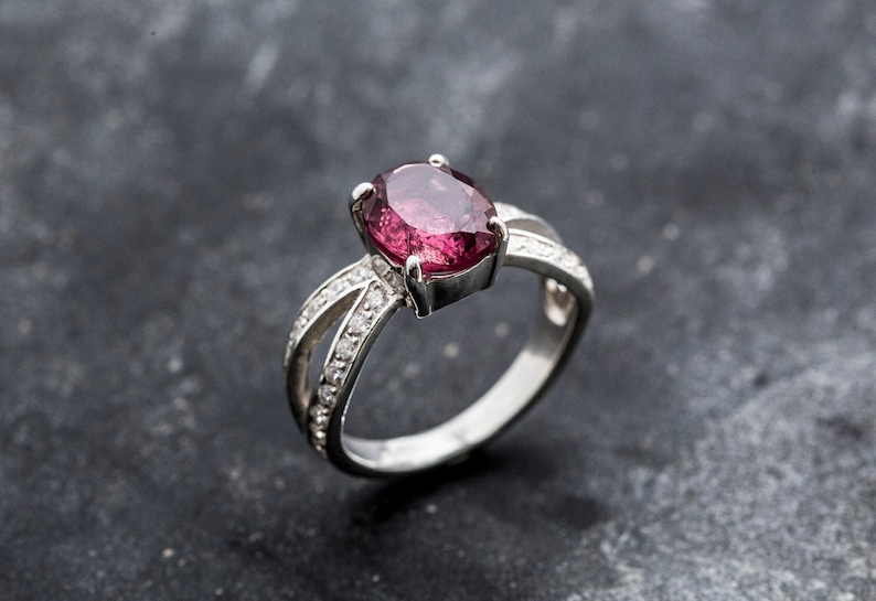 Pu&ograve; includere: Un anello d'argento con una grande gemma ovale rosa intenso. La fascia si divide in due su ogni lato, impreziosita da piccole gemme trasparenti. L'anello &egrave; posto su uno sfondo scuro e strutturato. Un gioiello.