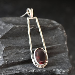 Puede incluir: Un colgante de plata con una piedra ovalada de color rojo oscuro engastada en un marco de plata trenzado.