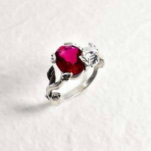 Puede incluir: Anillo de plata con una gema roja oscura de forma ovalada. El anillo presenta detalles de hojas y vides, creando un diseño inspirado en la naturaleza. La gema es el punto focal, engastada prominentemente en la intrincada filigrana.