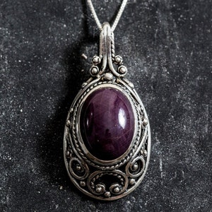 Puede incluir: Un collar con colgante de plata con una piedra preciosa ovalada de color p&uacute;rpura intenso. El colgante tiene un dise&ntilde;o de filigrana ornamentado y una fianza decorativa. El collar se muestra sobre una superficie oscura y texturizada.