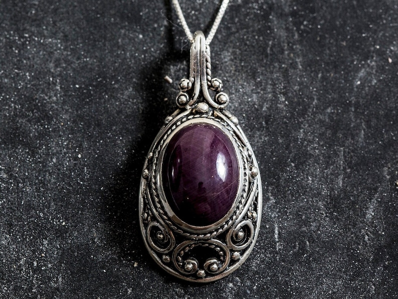 Puede incluir: Un collar con colgante de plata con una piedra preciosa ovalada de color p&uacute;rpura intenso. El colgante tiene un dise&ntilde;o de filigrana ornamentado y una fianza decorativa. El collar se muestra sobre una superficie oscura y texturizada.